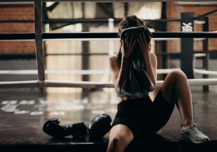 A female boxer sitting in a gym ring, resting and wiping sweat with a towel after a workout.